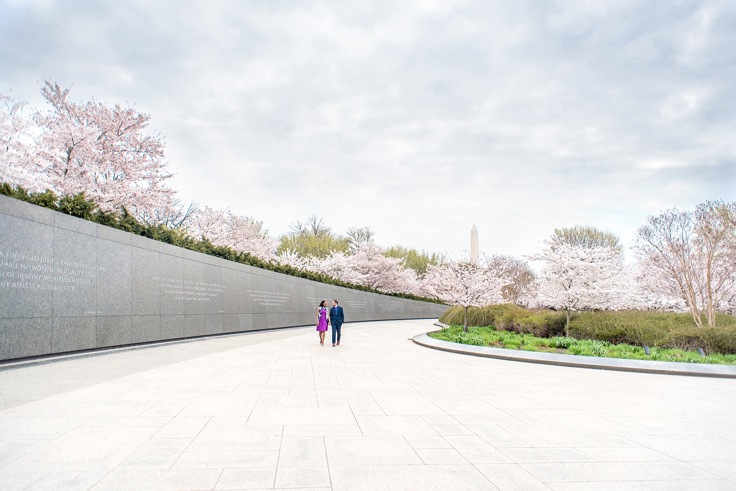 DC Cherry Blossoms Engagement Photos by Mikkel Paige Photography. Spring flowers at the Martin Luther King Jr. memorial with the Washington Monument in the background, at the nation's Capitol with an interracial couple in colorful outfits. #DCCherryBlossoms #CherryBlossoms #EngagementPhotos #SpringEngagementPhotos #CherryBlossomsEngagementPhotos