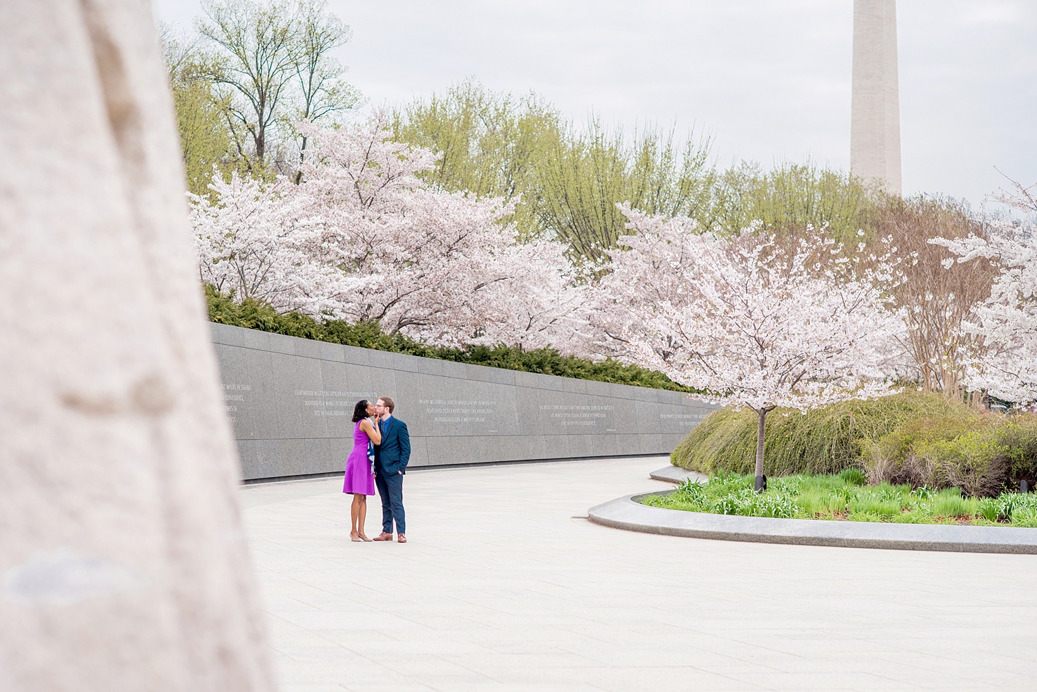 DC Cherry Blossoms Engagement Photos by Mikkel Paige Photography. Spring flowers at the Martin Luther King Jr. memorial with the Washington Monument in the background, at the nation's Capitol with an interracial couple in colorful outfits. #DCCherryBlossoms #CherryBlossoms #EngagementPhotos #SpringEngagementPhotos #CherryBlossomsEngagementPhotos