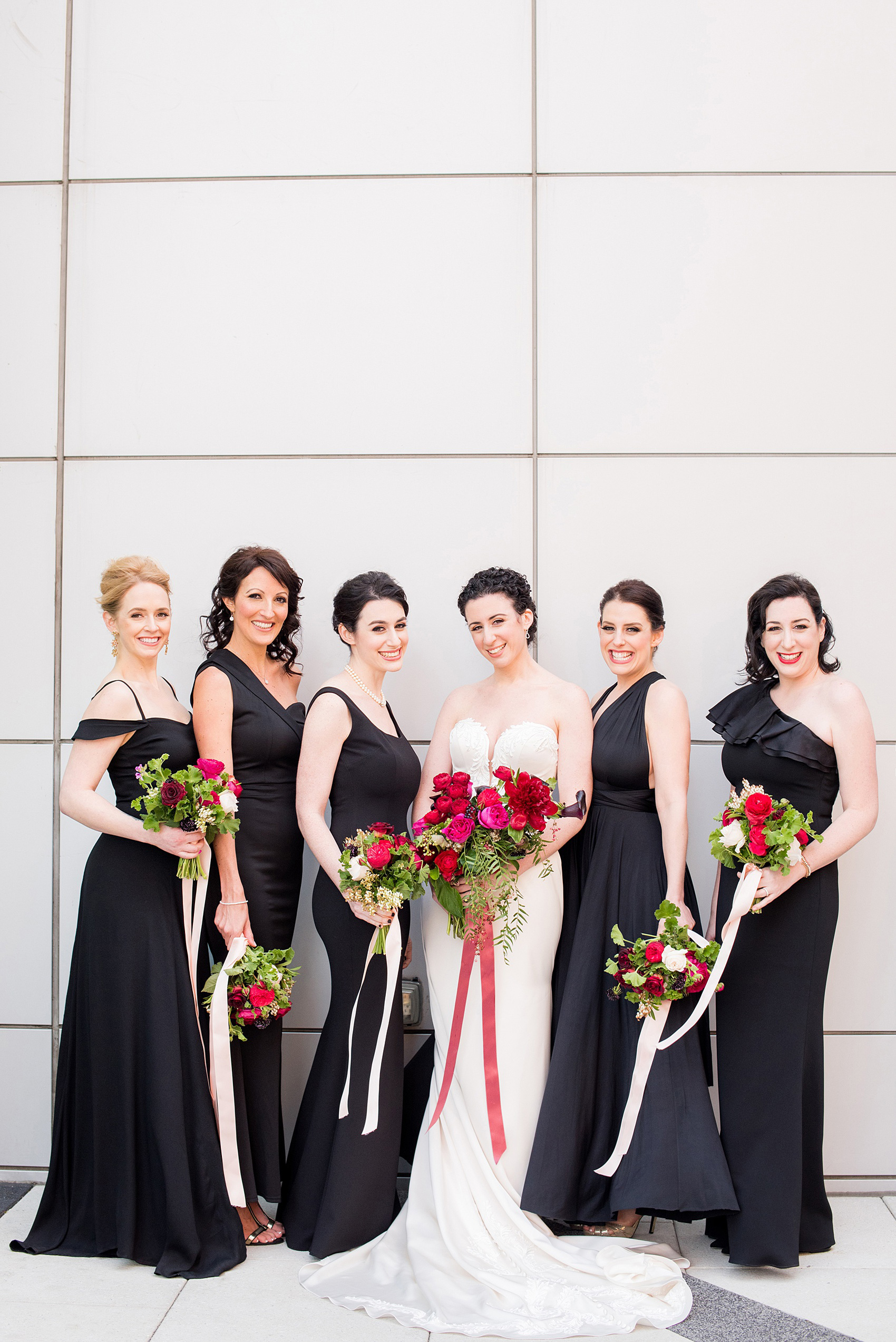 W Hoboken wedding photos at a New Jersey venue overlooking the NYC skyline. Picture by Mikkel Paige Photography of the bridal party in varied, mismatched black gowns carrying romantic bouquets in red and pink and long ribbons by Sachi Rose Designs. #mikkelpaige #HobokenWedding #NewJerseyPhotographer #NewYorkCityPhotographer #NYCweddingphotographer #brideandgroomphotos #redpeonies #romanticwedding #springwedding #CityWedding