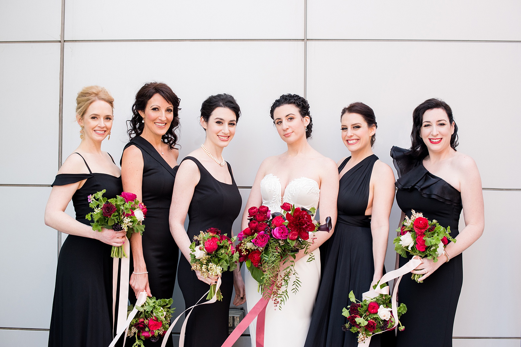 W Hoboken wedding photos at a New Jersey venue overlooking the NYC skyline. Picture by Mikkel Paige Photography of the bridal party in varied, mismatched black gowns carrying romantic bouquets in red and pink and long ribbons by Sachi Rose Designs. #mikkelpaige #HobokenWedding #NewJerseyPhotographer #NewYorkCityPhotographer #NYCweddingphotographer #brideandgroomphotos #redpeonies #romanticwedding #springwedding #CityWedding