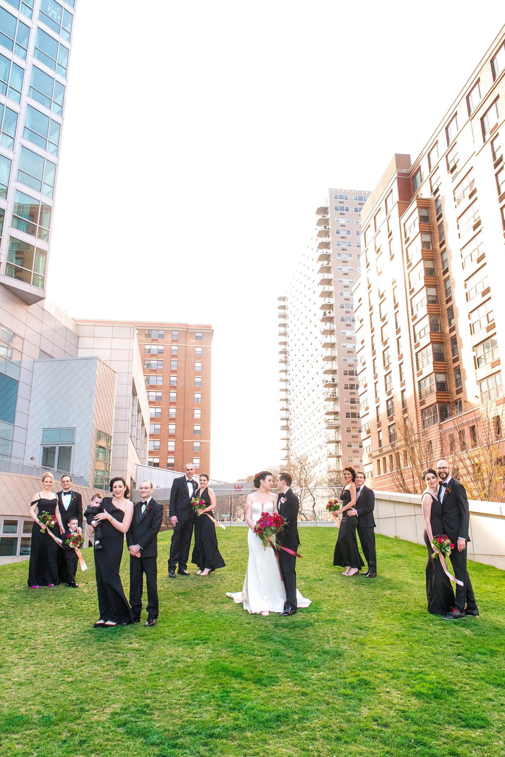 W Hoboken wedding photos at a New Jersey venue overlooking the NYC skyline. Vogue like photo by Mikkel Paige Photography of the bridal party in black gowns carrying romantic bouquets in red and pink and long ribbons blowing in the breeze and groomsmen in classic tuxedos for a black tie wedding. #mikkelpaige #HobokenWedding #NewJerseyPhotographer #NewYorkCityPhotographer #NYCweddingphotographer #brideandgroomphotos #redpeonies #romanticwedding #springwedding #CityWedding