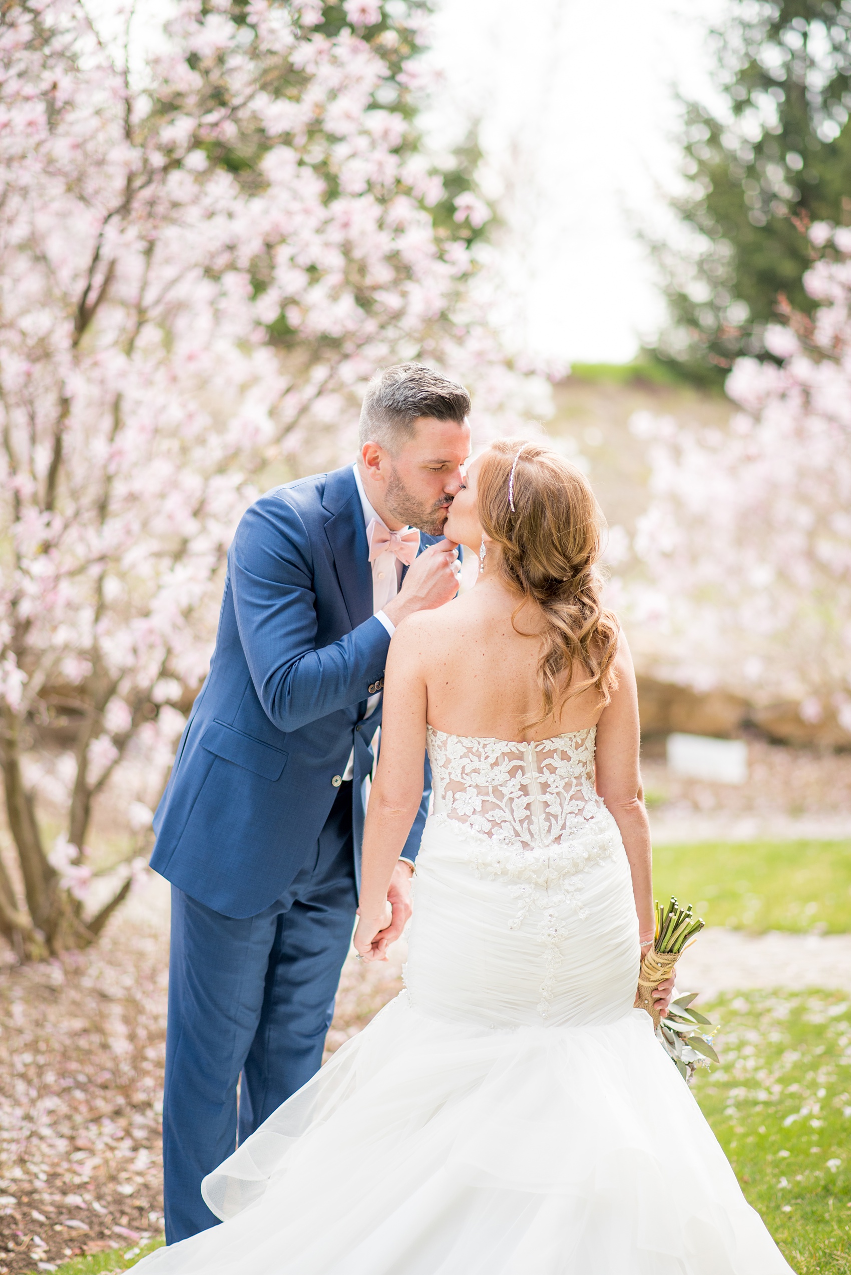 Crystal Springs Resort wedding photos in New Jersey, with photographer Mikkel Paige Photography. The couple’s spring wedding had an outdoor ceremony with photos at this rustic, woodsy venue showing their blue and pink palette decor from getting ready to their reception. This picture shows the groom in a custom navy blue suit and pink bow tie gently kissing his bride, in her lace gown, amongst the cherry blossoms. Click through to see their complete wedding recap! #NewJerseywedding #MikkelPaige #NJweddingphotographer #NJphotographer #brideandgroom #springwedding