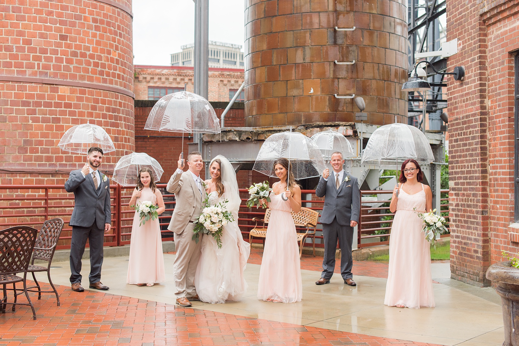 The Rickhouse wedding photos by Mikkel Paige photography in Durham, North Carolina. This urban venue has all the charm of the south for this bride and groom's spring wedding! This picture shows the bridal party posing for a photo in the rain with clear umbrellas at American Tobacco Campus. Click to see more from their day! #mikkelpaige #northcarolina #southernwedding #TheRickhouseDurham #rosegoldandpink #rainywedding #weddingintherain #bridalparty #umbrellabridalparty