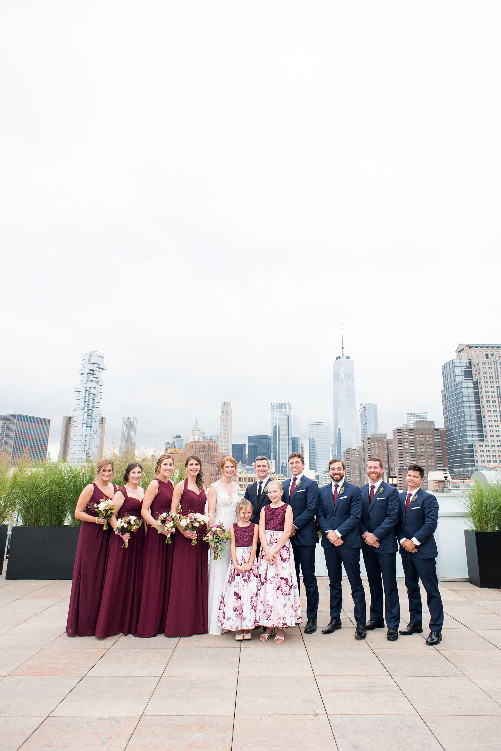 Photos of a NYC wedding venue, Tribeca Rooftop, by Mikkel Paige Photography. This ceremony and reception location has an outdoor space overlooking the Manhattan skyline. These pictures will provide inspiration in New York! The bride and groom were surrounded by bridesmaids in cranberry gowns and groomsmen in navy blue suits, holding fall flowers. Click through for more details from this celebration! #TribecaRooftop #MikkelPaige #NYCwedding #NYCWeddingVenues #NYCWeddingPhotography #FallFlowers
