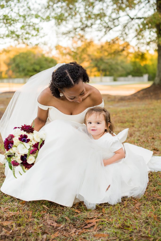 A fall wedding with burgundy, dusty rose and grey details. The bride wore an off-the-shoulder Anne Barge dress and knelt down to talk to her niece, the flower girl. Mikkel Paige Photography, photographer in Greenville NC and Raleigh captured this wedding at Rock Springs Center, planned by @vivalevent. Click through for more details and pictures from this autumn day! #mikkelpaige #northcarolinawedding #southernwedding #burgundywedding #offtheshoulder #annebarge #flowergirl #bridestyle