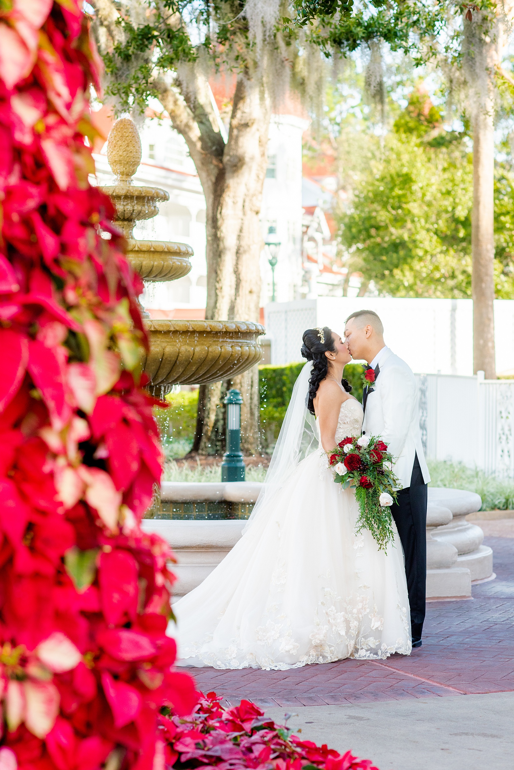 Walt Disney World photographs by Mikkel Paige Photography. The bride and groom had December pictures at the Grand Floridian Resort, Wedding Pavilion and Contemporary Hotel. It had a Beauty and the Beast theme and the bride wore a beautiful rose embroidered dress, befitting of a princess. Her red rose bouquet had holiday greenery and a cascading effect. The groom wore a white tuxedo. #disneywedding #disneybride #waltdisneyworld #DisneyWorldWedding #cascadingbouquet