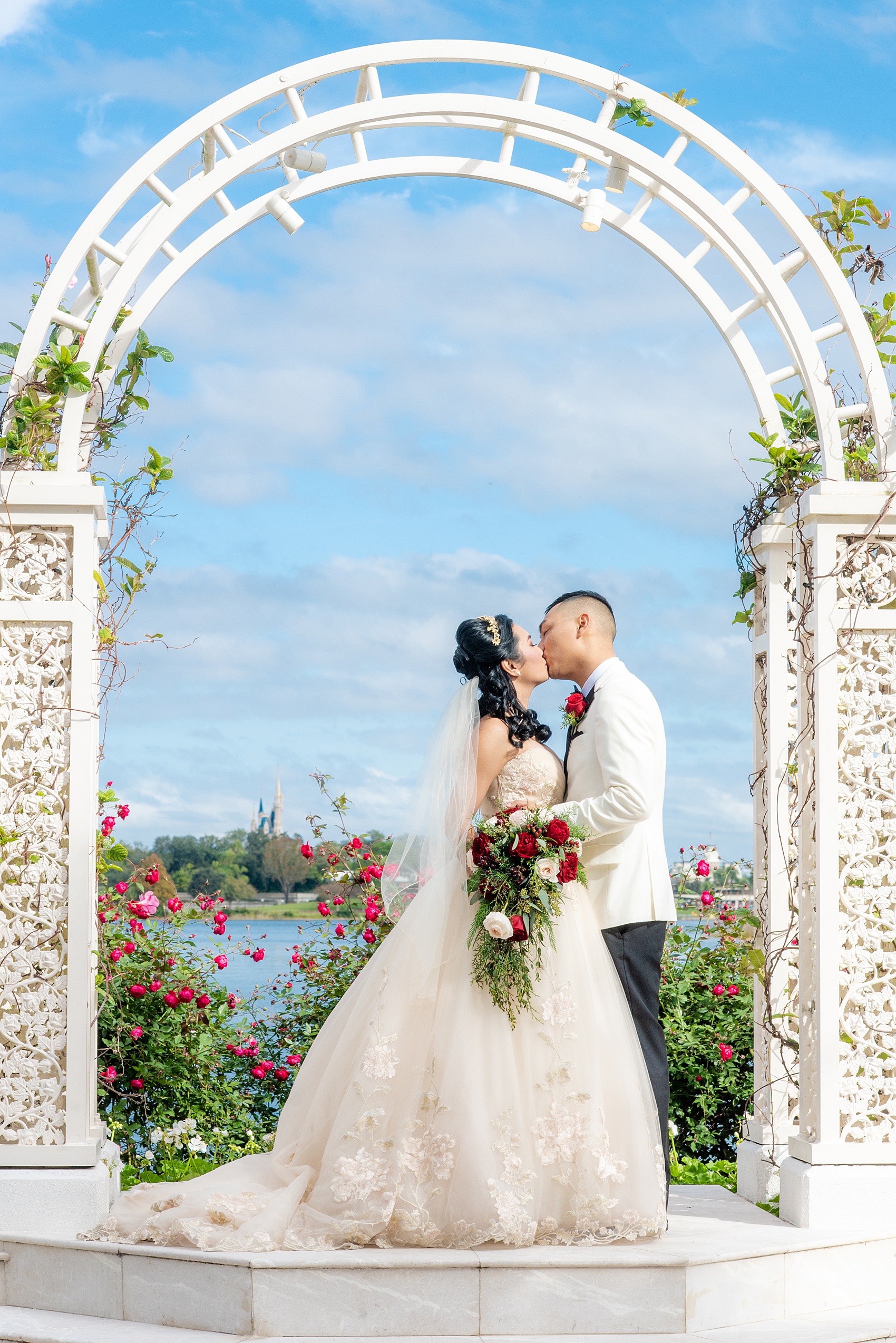 Walt Disney World photographs by Mikkel Paige Photography. The bride and groom had December pictures at the Grand Floridian Resort, Wedding Pavilion and Contemporary Hotel. It had a Beauty and the Beast theme and the bride wore a beautiful rose embroidered dress, befitting of a princess. Her red rose bouquet had holiday greenery and a cascading effect. The groom wore a white tuxedo. #disneywedding #disneybride #waltdisneyworld #DisneyWorldWedding #cascadingbouquet