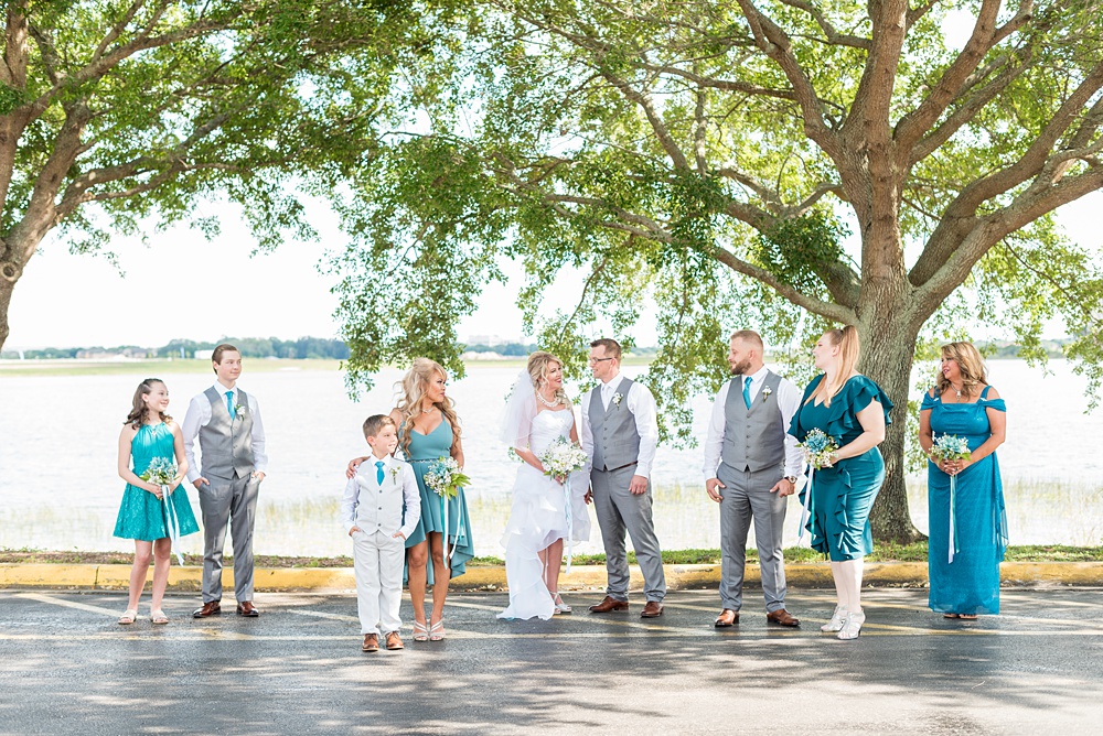 Hub 925 wedding photos at an alternative Orlando wedding venue by Mikkel Paige Photography. The bride, groom and their wedding party posed for a photo by Sand Lake. The bridesmaids wore different shades of teal dresses and groomsmen wore grey slacks and vests. #mikkelpaige #hub925 #orlandoweddingphotographer #orlandoweddingphotos #floridaweddingphotographer #centralfloridawedding #bridalparty #tealbridesmaids #weddingparty