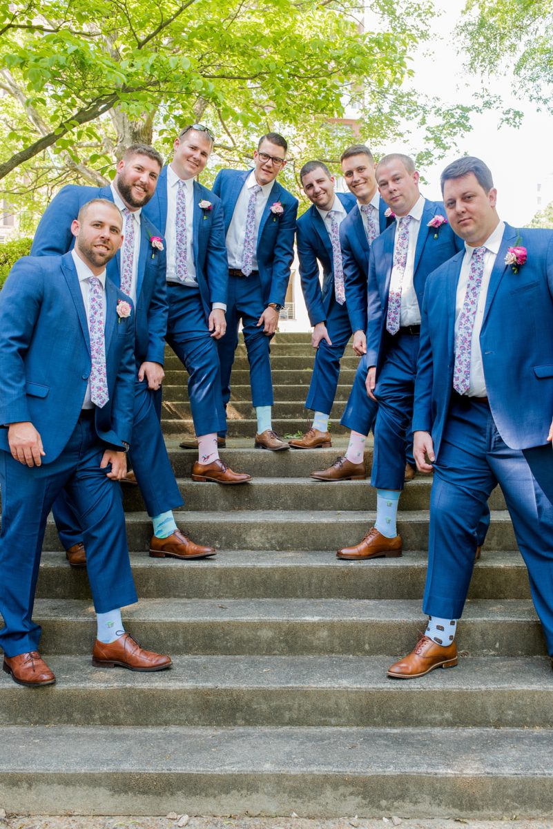 Raleigh wedding photographer, Mikkel Paige Photography, pictures of the groom and his groomsmen in blue suits with floral ties in downtown Raleigh, North Carolina at the event venue The Stockroom at 230 and capital building. The bright colors and hot pink boutonnieres were perfect for a spring May celebration. #MikkelPaige #DowntownRaleigh #RaleighWedding #RaleighVenue #TheStockroomat230 #capitalcity #groomsmen #boutonnieres #bluesuits #floralties #groomstyle