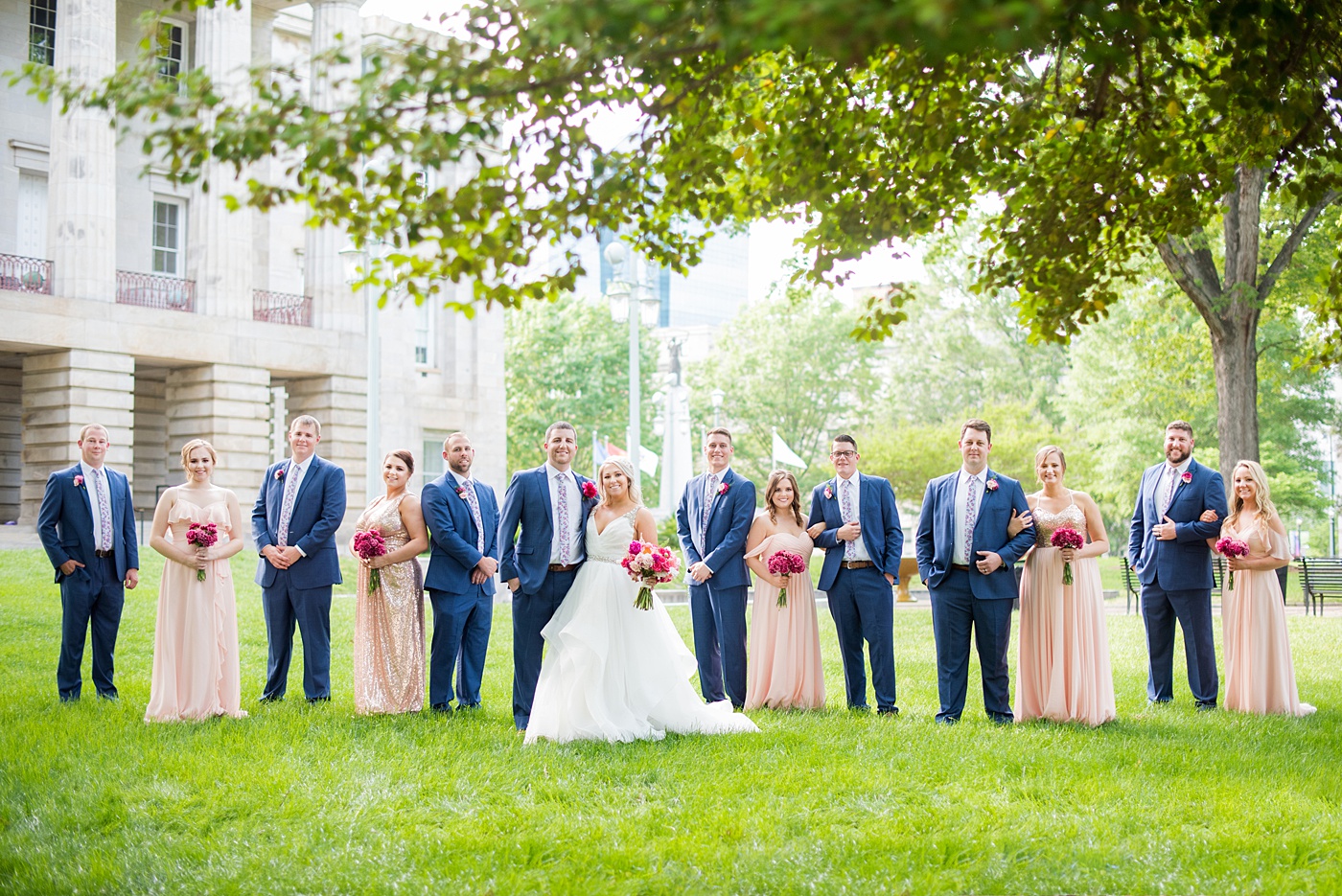 Raleigh wedding photographer, Mikkel Paige Photography, pictures of mismatched pink bridesmaids + groomsmen in blue suits with floral ties in downtown Raleigh, North Carolina at the event venue The Stockroom at 230, The Glass Box, and capital building. Their hot pink peony + carnation bouquets were perfect for a spring May celebration. #MikkelPaige #DowntownRaleigh #RaleighWedding #RaleighVenue #TheStockroomat230 #capitalcity #peonies #carnations #pinkbridesmaids #bridalpartyphotos #weddingparty