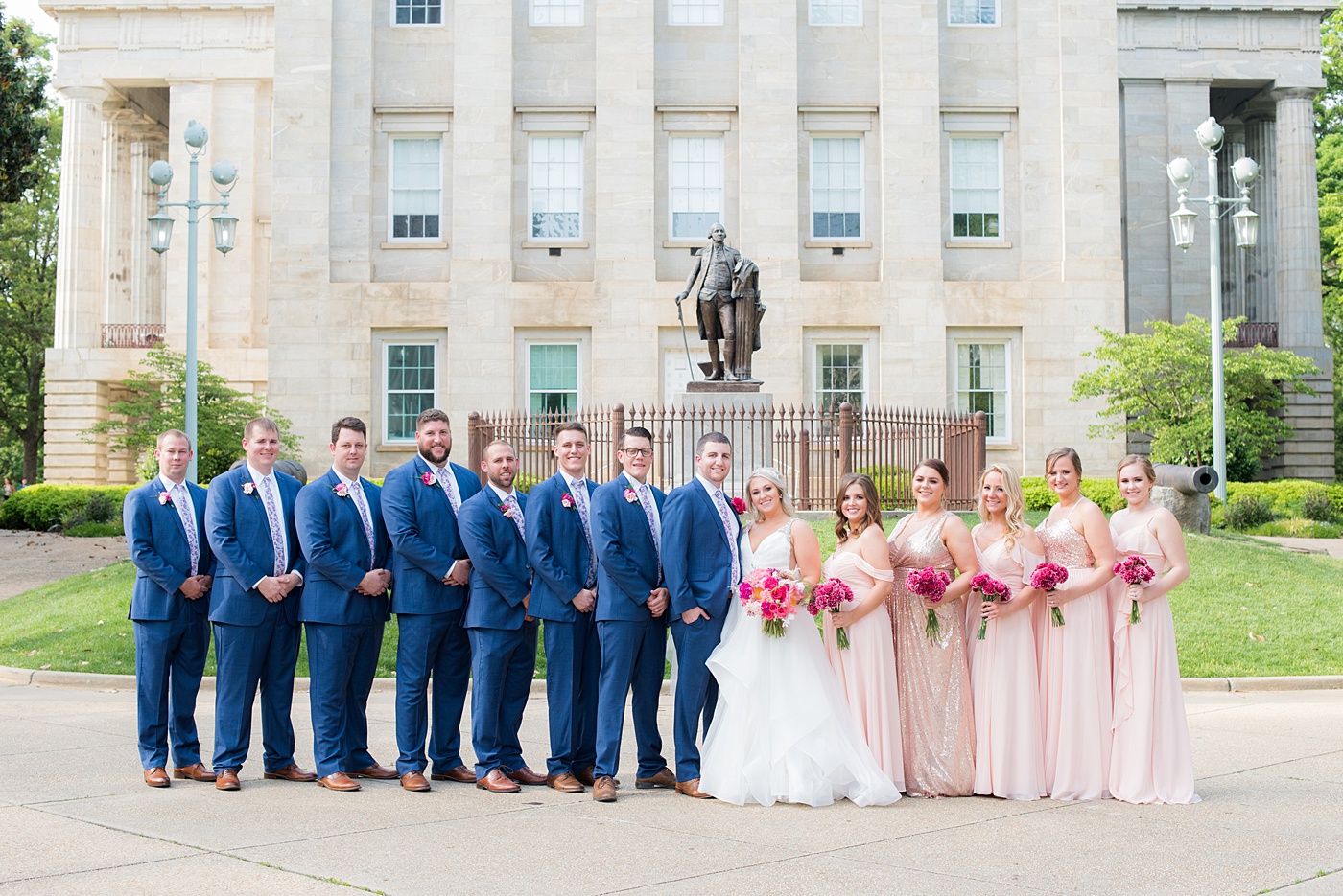 Raleigh wedding photographer, Mikkel Paige Photography, pictures of mismatched pink bridesmaids + groomsmen in blue suits with floral ties in downtown Raleigh, North Carolina at the event venue The Stockroom at 230, The Glass Box, and capital building. Their hot pink peony + carnation bouquets were perfect for a spring May celebration. #MikkelPaige #DowntownRaleigh #RaleighWedding #RaleighVenue #TheStockroomat230 #capitalcity #peonies #carnations #pinkbridesmaids #bridalpartyphotos #weddingparty