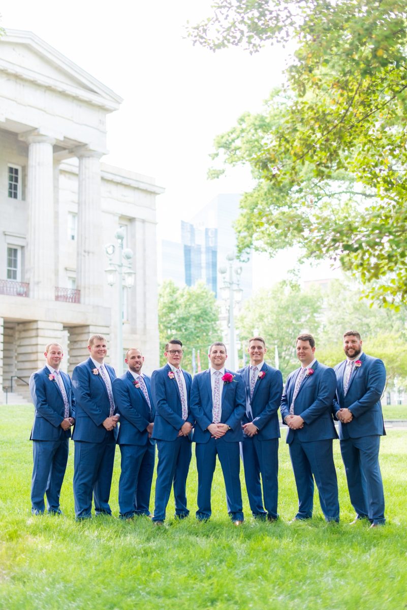 Raleigh wedding photographer, Mikkel Paige Photography, pictures of the groom and his groomsmen in blue suits with floral ties in downtown Raleigh, North Carolina at the event venue The Stockroom at 230 and capital building. The bright colors and hot pink boutonnieres were perfect for a spring May celebration. #MikkelPaige #DowntownRaleigh #RaleighWedding #RaleighVenue #TheStockroomat230 #capitalcity #groomsmen #boutonnieres #bluesuits #floraties #groomstyle #layflatphotography