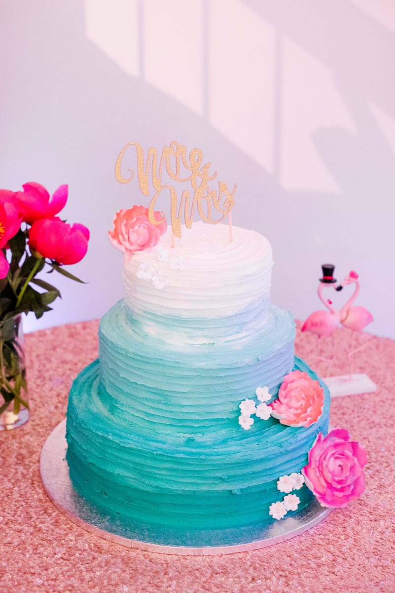 A spring wedding in downtown Raleigh, North Carolina, at the event venue The Stockroom at 230 with a Publix cake in ombre blue with a “Mr. and Mrs.” cake topper and pink flamingos nearby. Mikkel Paige Photography, their photographer, captured a lay flat of their menu from the reception, with hot pink and aqua blue watercolors and flower detail. #MikkelPaige #DowntownRaleigh #RaleighWedding #RaleighVenue #TheStockroomat230 #weddingreception #weddingmenu #weddingcake #ombrecake