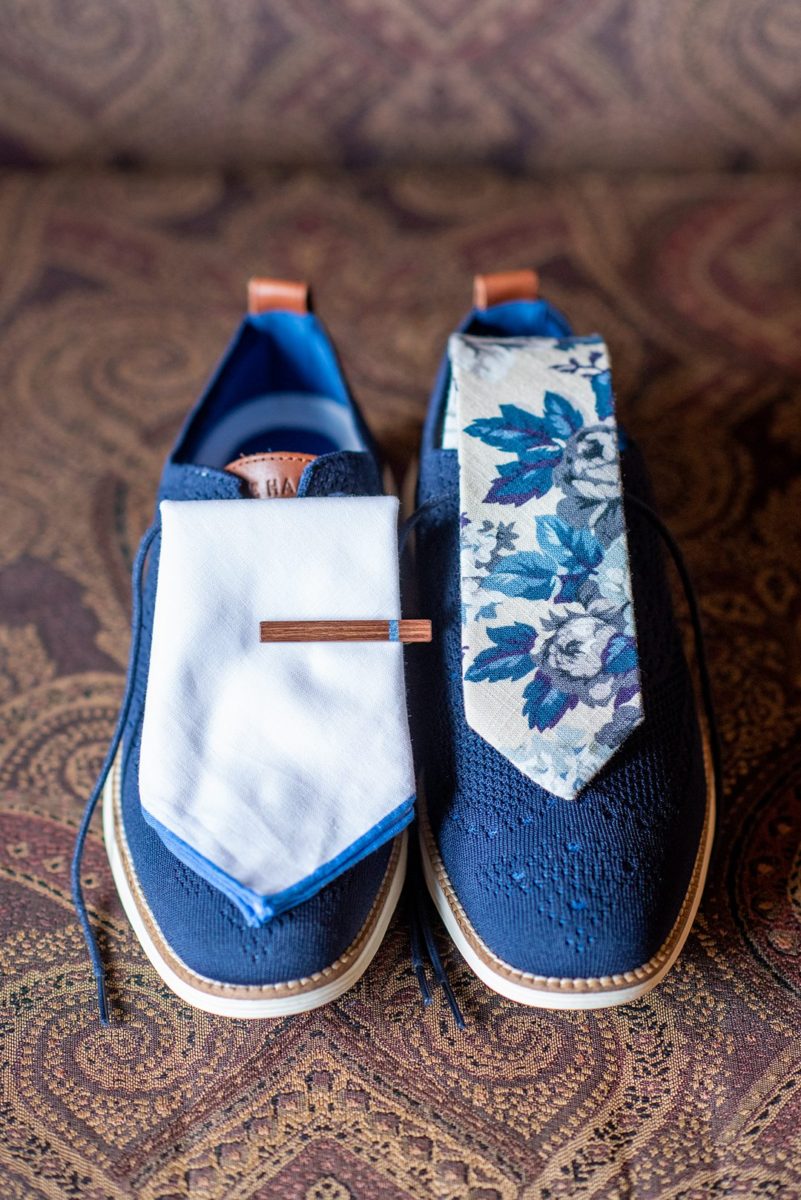 Photos at New Jersey wedding venue, Crystal Springs Resort, in Hamburg with an outdoor ceremony option and indoor reception, by Mikkel Paige Photography. The groom got ready for his day with his groomsmen and father-in-law and wore a blue suit. This detail picture shows his blue wedding shoes, pocket square and wooden tie clip, and floral tie. #mikkelpaige #CrystalSprings #NJweddingvenues #NewJerseyWedding #NJweddingphotographer #bluesuit #groomstyle #gettingready #groomshoes