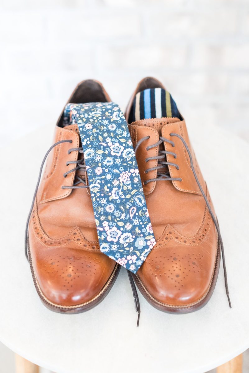 Detail photo of the groom's floral blue tie and brown leather shoes and striped socks at Chatham Station by Mikkel Paige Photography. This beautiful Cary, North Carolina venue has indoor and outdoor spaces. #mikkelpaige #RaleighWeddingPhotographer #NorthCarolinaWeddings #SouthernWeddings #groomstyle #gettingready #floraltie #flowertie