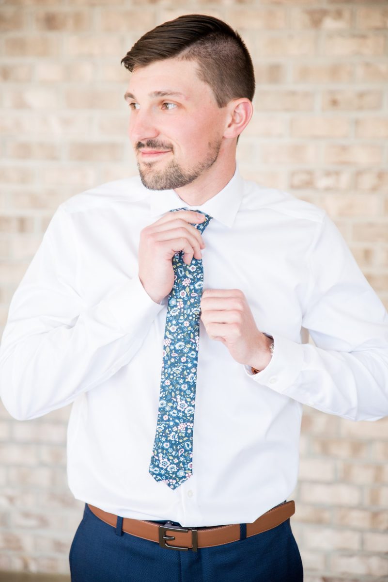 Detail photo of the groom tying his floral blue tie for his custom jacket at Chatham Station by Mikkel Paige Photography. This beautiful Cary, North Carolina venue has indoor and outdoor spaces. #mikkelpaige #RaleighWeddingPhotographer #NorthCarolinaWeddings #SouthernWeddings #groomstyle #customsuit #weddingdetails #gettingready #floraltie #flowertie