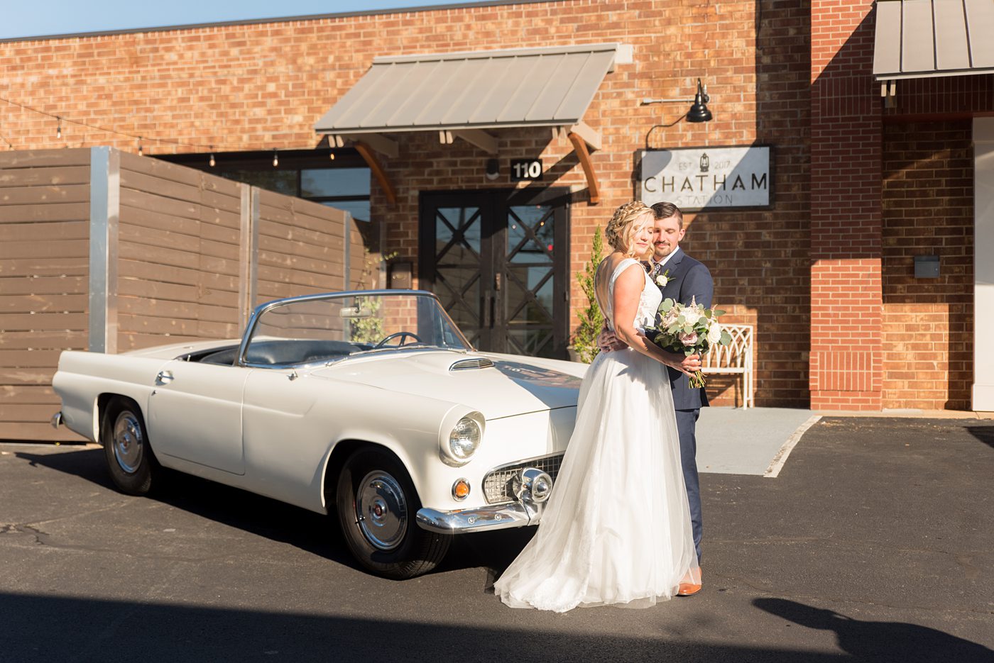 Chatham Station wedding photos by Mikkel Paige Photography from this urban chic beautiful venue in Cary, North Carolina. The bride wore a tulle v-neck gown and up hair-do with braids and groom a blue suit with custom details for a fall celebration. They took iconic photos with a vintage white Thunderbird car. #mikkelpaige #RaleighWeddingPhotographer #NorthCarolinaWeddings #SouthernWeddings #bridestyle #brideandgroom #vintagecar