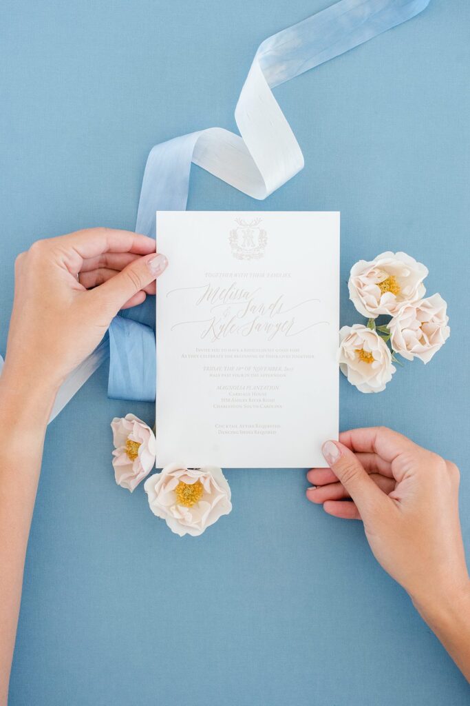 Hands holding the corners of an elegant invitation with a blue ribbon behind it and rose heads.