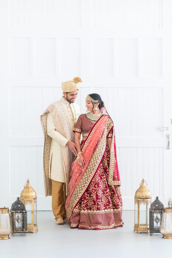 Bride and groom in Indian wedding attire in front of a white wall with decorative metal lanterns surrounding them at The Graham Mill wedding venue in NC. Photos by Mikkel Paige Photography.