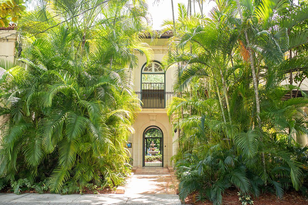 Front entrance archway and second floor Juliet balcony framed by palm trees, at Villa Woodbine. This historic home is a wedding venue in Miami, FL. Picture by Mikkel Paige Photography.