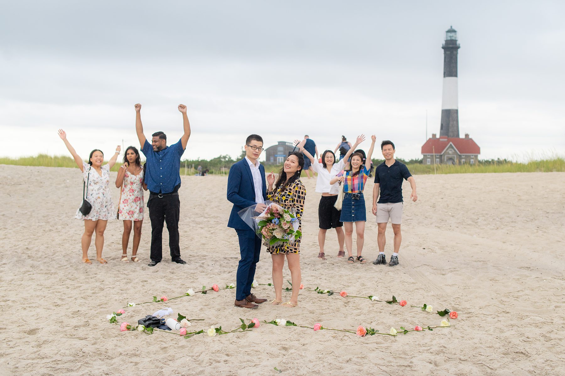 Photo of a couple in dress clothes with the Fire Island Lighthouse behind them after a proposal at the beach on Long Island with a heart made from rose stems in the sand, and friends nearby.