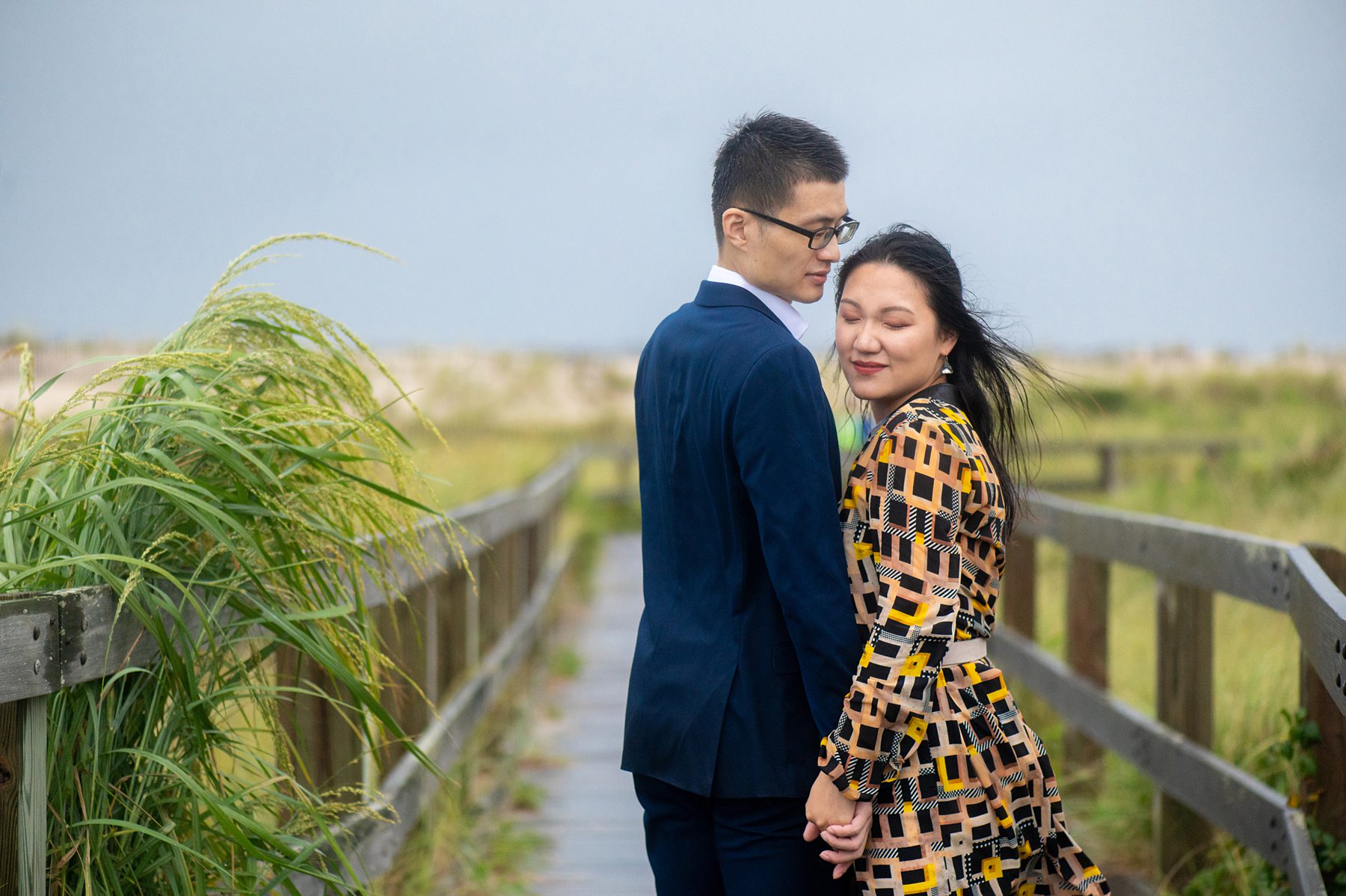 Photo of a couple at the beach with greenery blurred behind them as they walk on a boardwalk on Long Island.