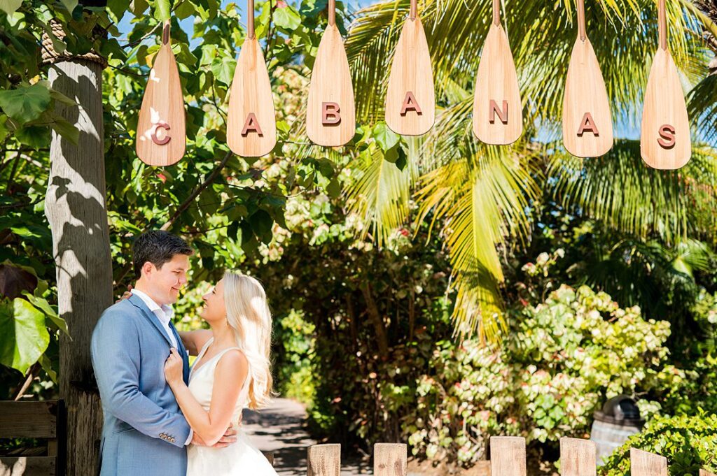 Bride and groom under the Cabanas sign with lush tropical foliage behind them on Castaway Cay for wedding photos. Pictures by Mikkel Paige Photography.