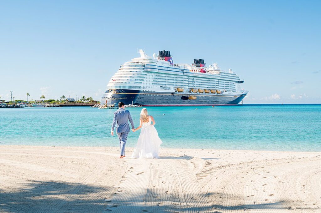 Bride and groom walking on the beach with Disney Wish cruise ship in the background on Castaway Cay. Images by Mikkel Paige Photography.
