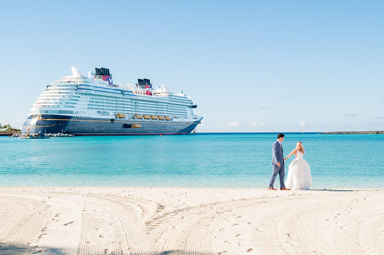 Bride and groom walking on the beach with Disney Wish cruise ship in the background on Castaway Cay. Images by Mikkel Paige Photography.