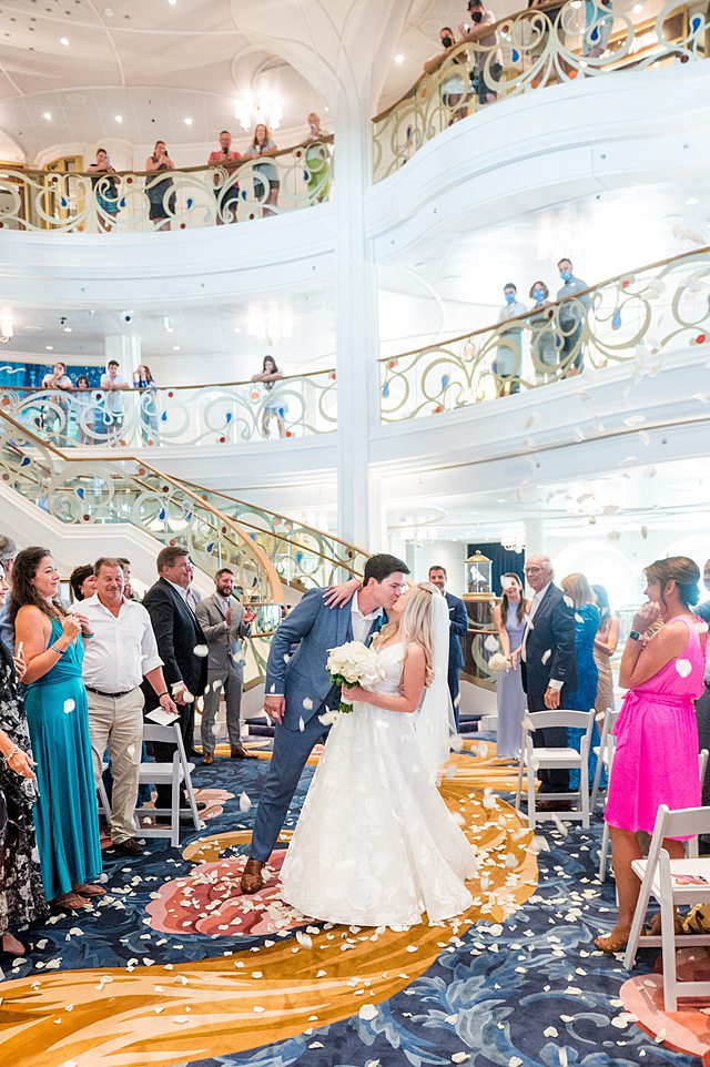 Couple kissing during a rose petal toss during their wedding ceremony in the grand hall on Disney Wish. Photos by Mikkel Paige Photography.