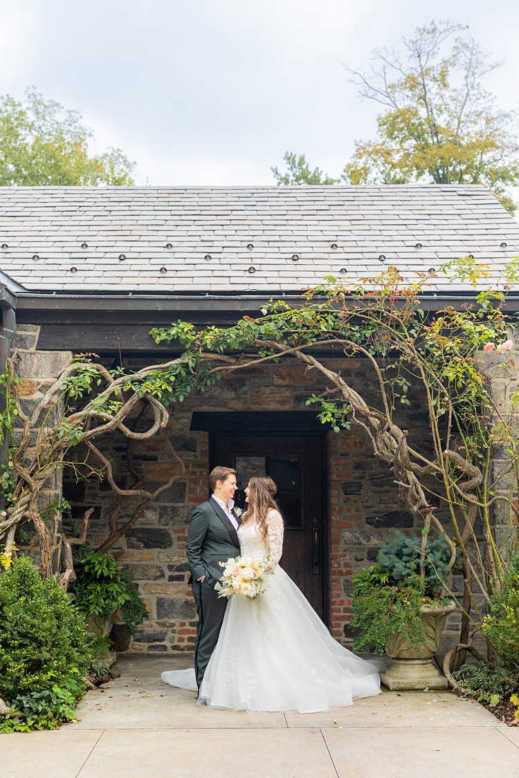 Two brides under a natural arch of branches in front of a building at their wedding at Blue Hill Stone Barns.