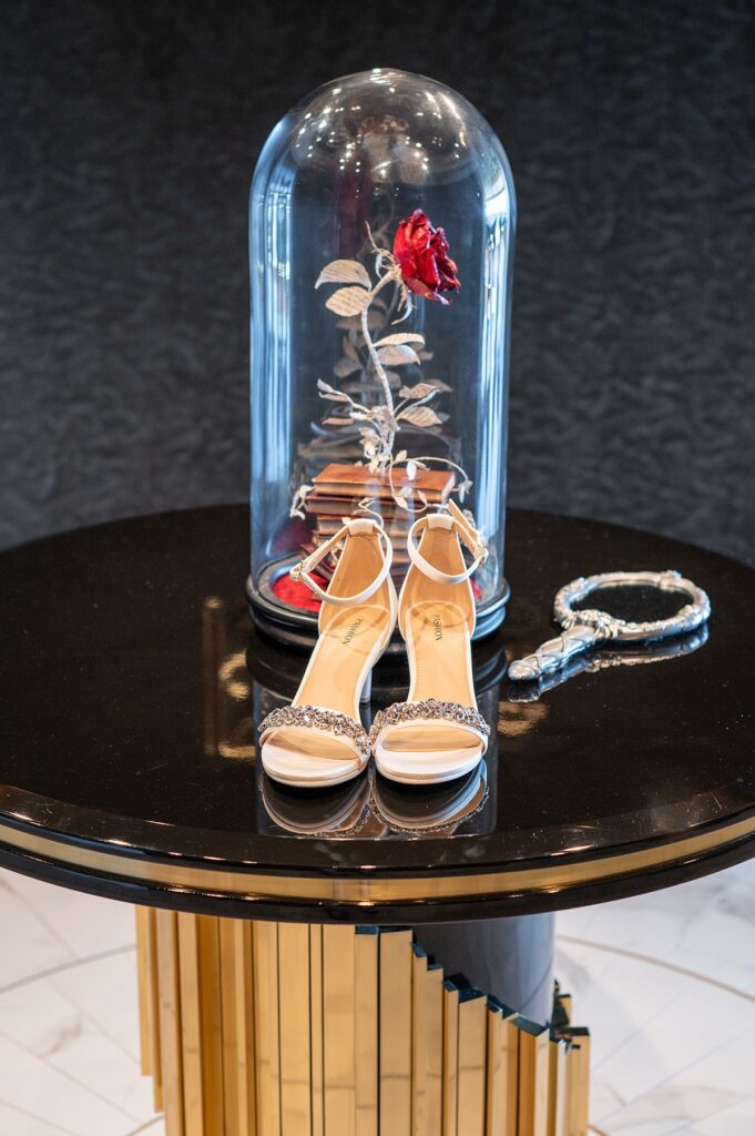 Wedding shoes on a table with a rose under a glass dome and a hand mirror next to it.